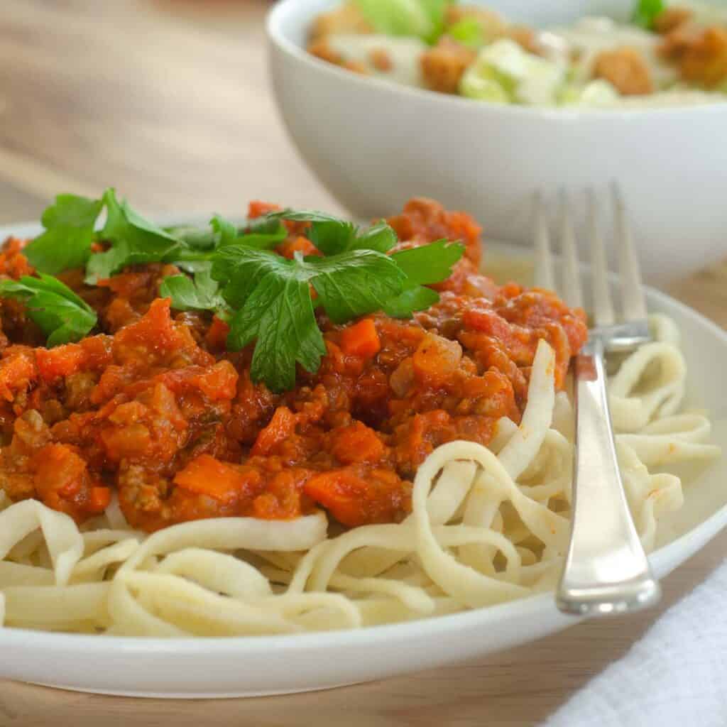 up close picture of bolognese sauce on top of a bed of linguine and a caesar salad in the background. 