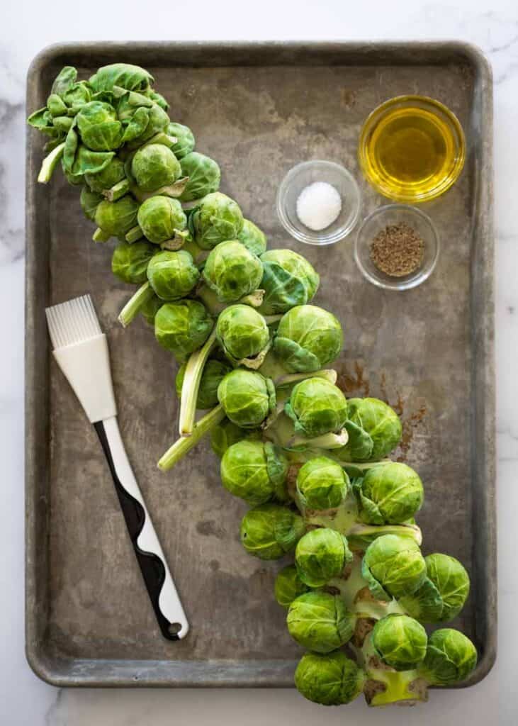 baking tray with Brussels sprouts stalk with bowls of olive, salt, pepper, and a pastry brush. 