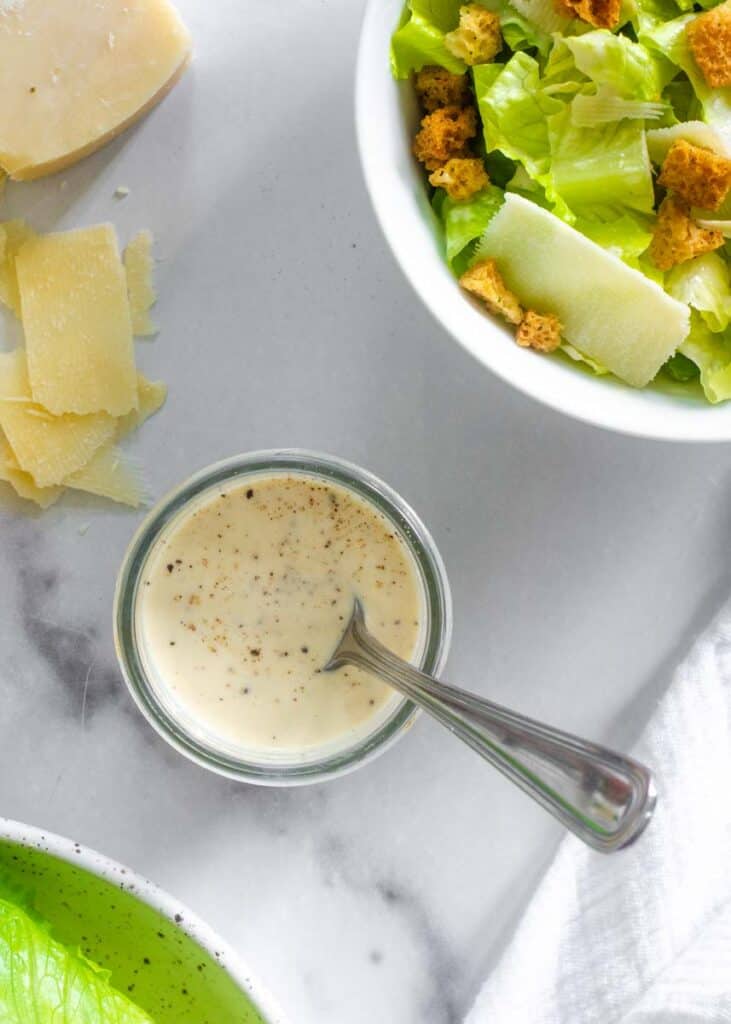 aerial shot of small jar of Caesar salad dressing next to a big bowl of salad. 