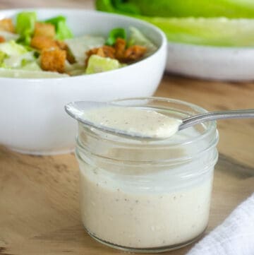 glass jar of caesar salad dressing with a spoon resting on top and a salad in the background.