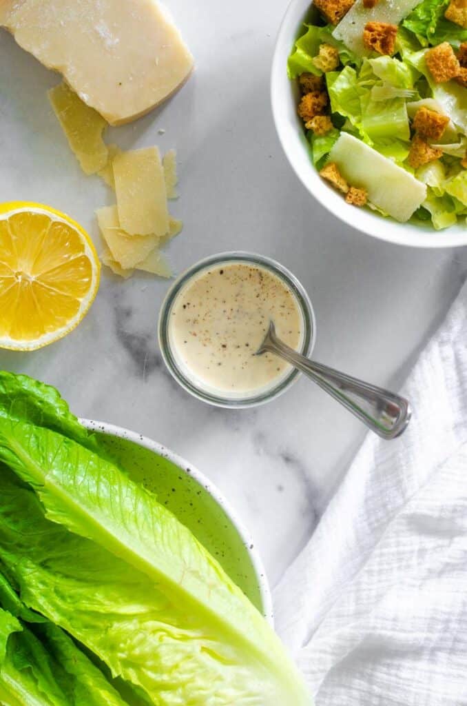 flat lay picture of a small jar of caesar salad dressing next to romaine hearts, sliced lemon, and a big bowl of salad. 