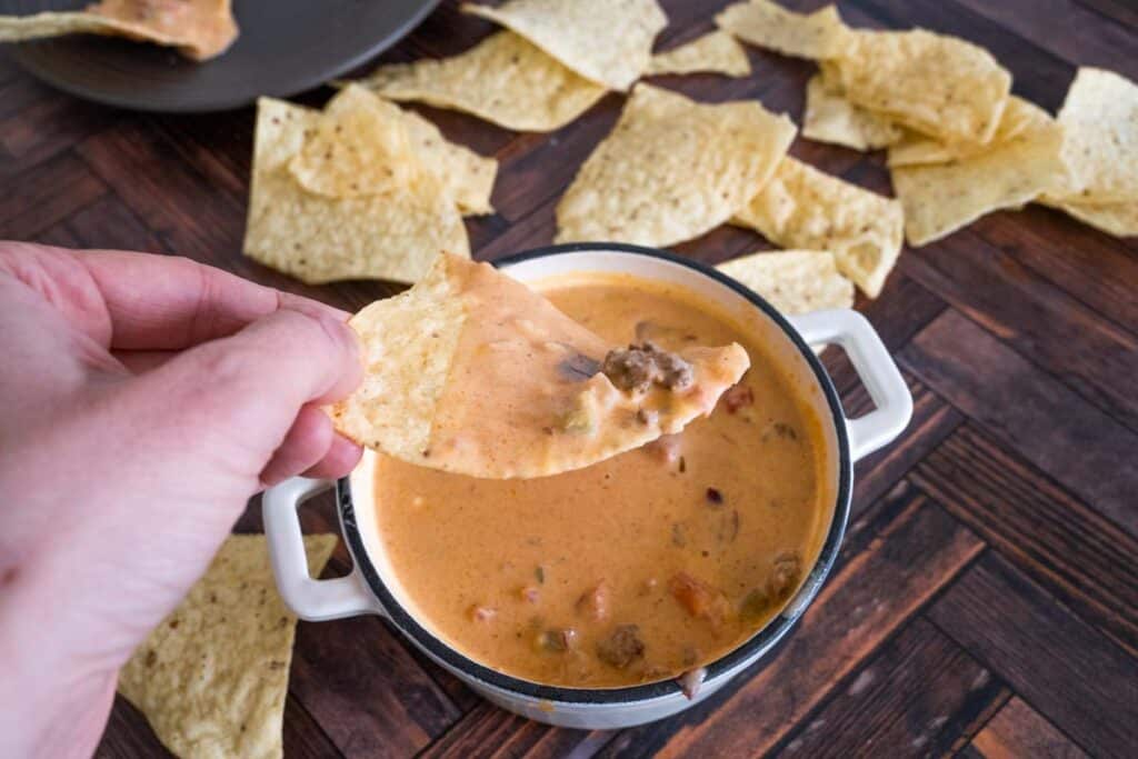 horizontal picture of a hand grabbing a chip with chili cheese dip on it.