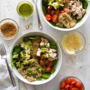 two white bowls with salad and smaller bowls of tomatoes, cheese, breadcrumbs, and dressing are scattered around the two bowls.