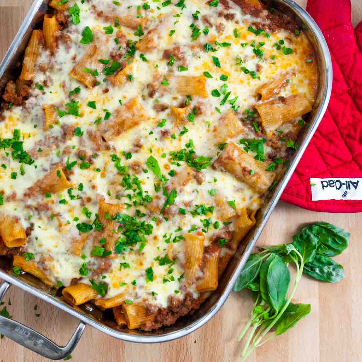 aerial photo of baked rigatoni al forno in large baking dish next to fresh basil and red oven mitt. 