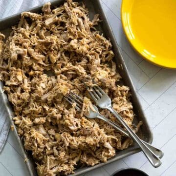 tray full of shredded chicken with two forks crossed on top, next to a yellow plate and small bowl of BBQ sauce.