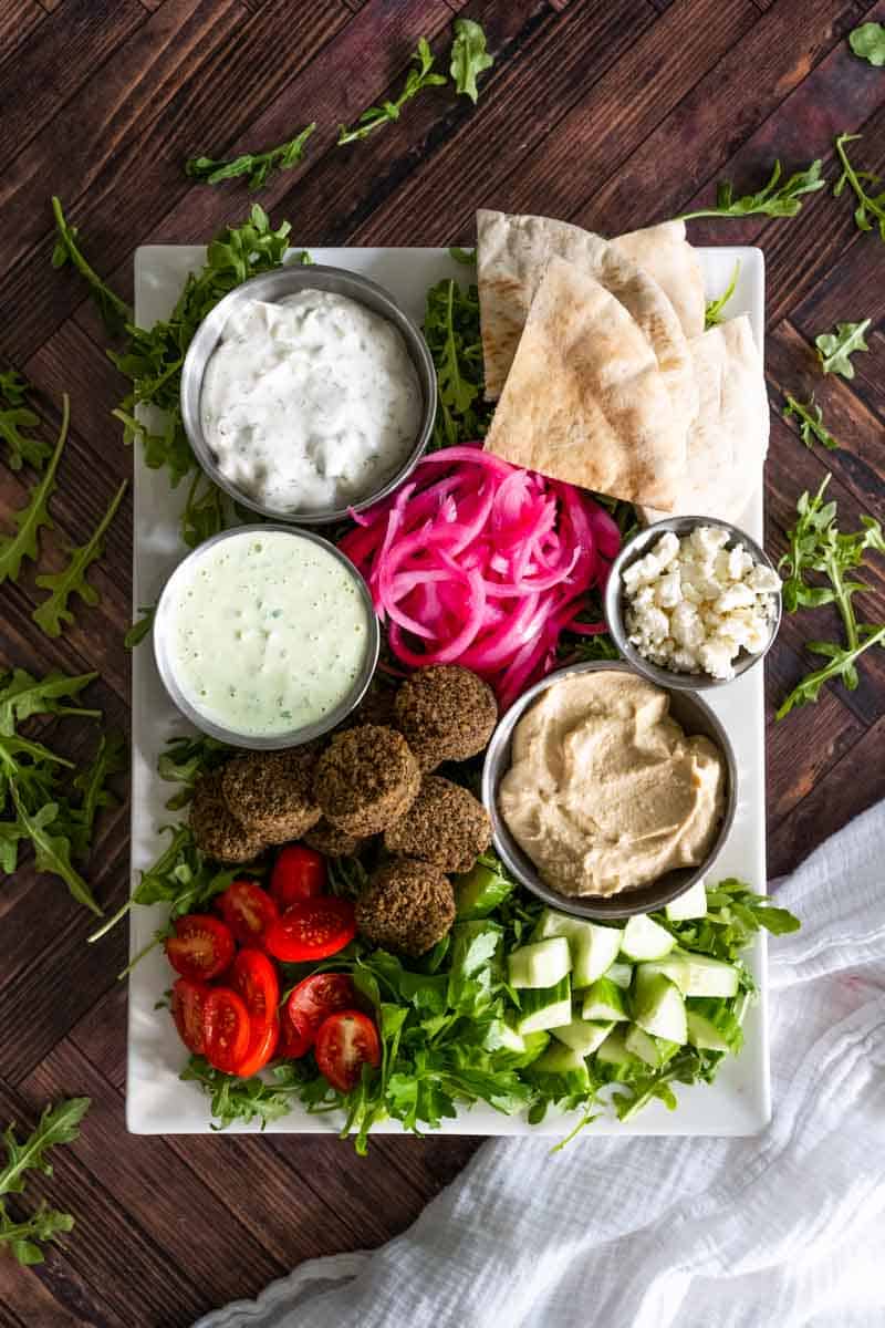 falafel on a white rectangular platter with a variety of ingredients in different sized bowls. 