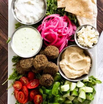 platter full of falafel, pickled onions, cucumbers, tomatoes, different dips and sauces all on a bed of arugula.