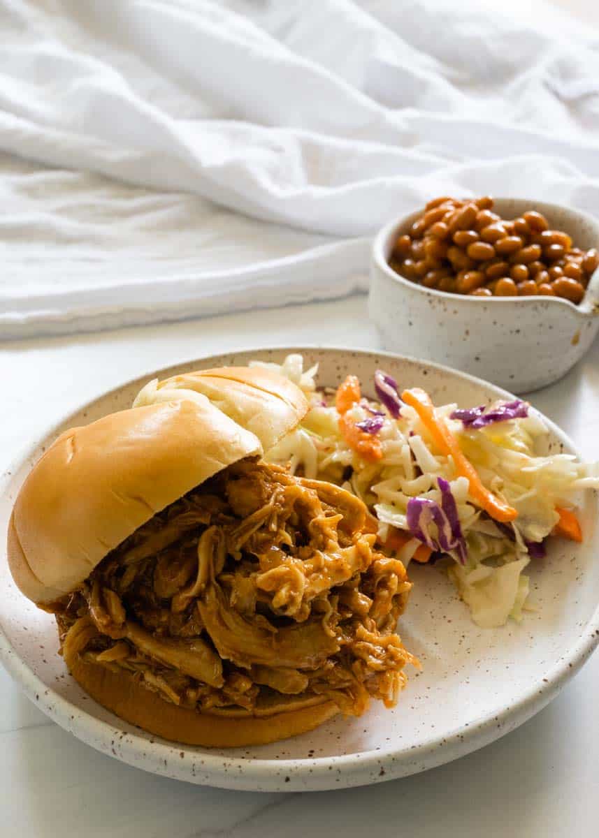 BBQ pulled chicken burger on a plate with cole slaw and baked beans in the background. 