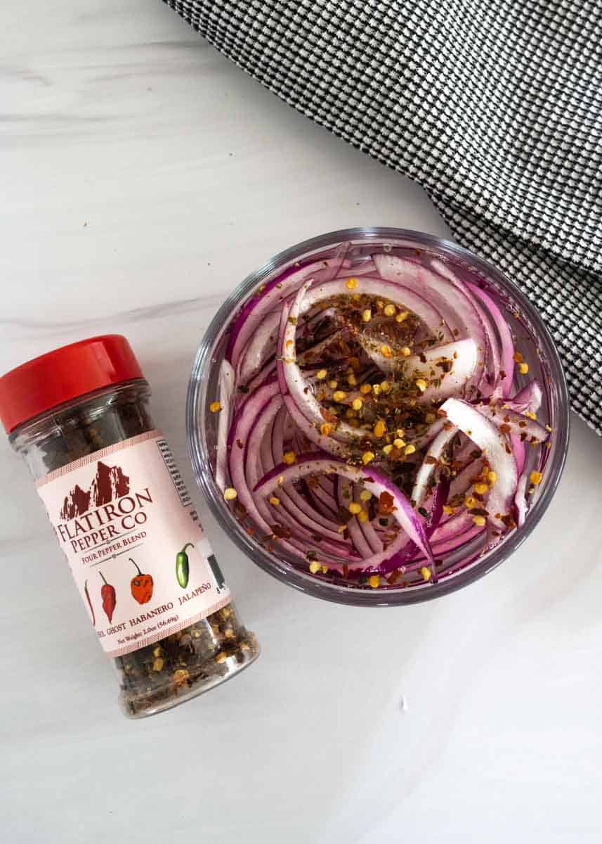 Container of freshly cut red onions in a vinegar mixture with a hot pepper blend poured on top. 