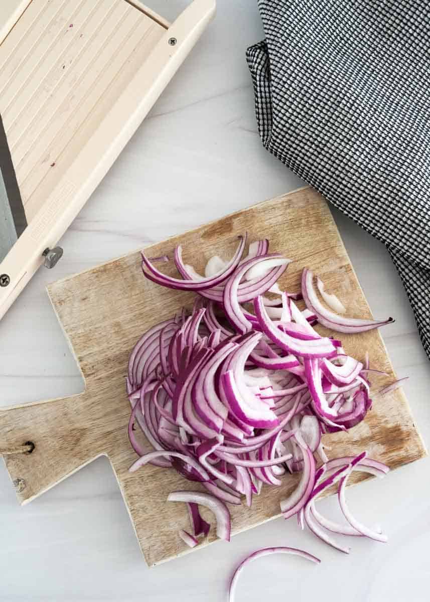 Thinly sliced red onions on wood cutting board next to a mandolin. 