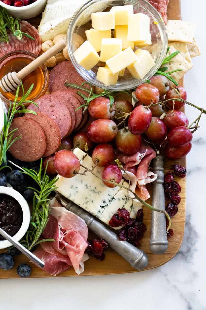 close up of a corner of a holiday board, with grapes, sausage, blue cheese, prosciutto, cheddar, blueberries, and dried cranberries. 