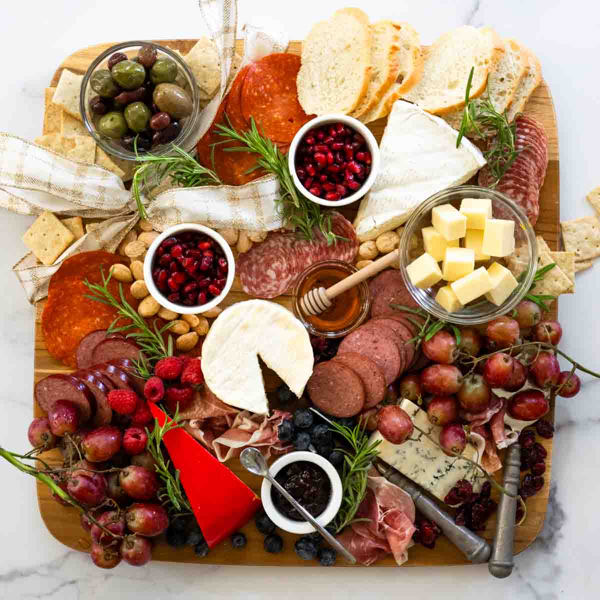 flatlay of a wooden board on marble backdrop filled with meats, cheeses, fruits, vegetables, and fresh rosemary sprigs. 