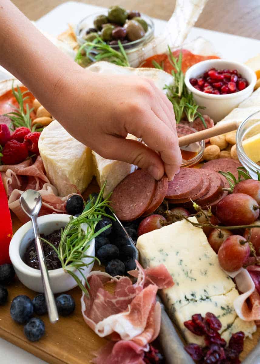 a small child's hand reaching for a slice of smoked sausage from a packed charcuterie board. 