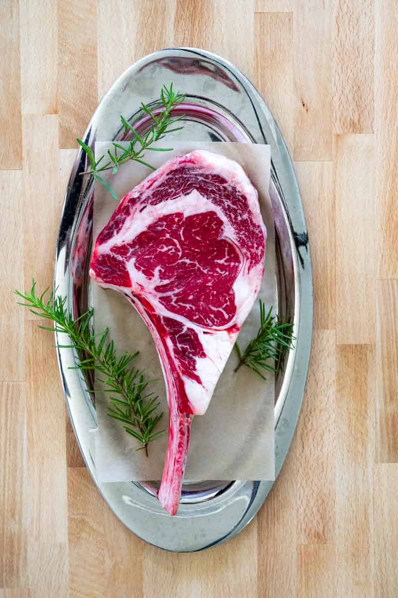 flatlay picture of a raw, thick tomahawk steak with long bone in on silver platter on top of butcher board. 