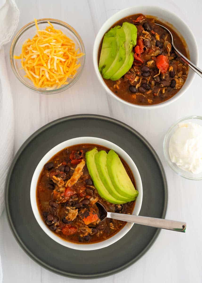 Two bowls of chili in white bowls with spoons stuck in them all over a larger black plate. 