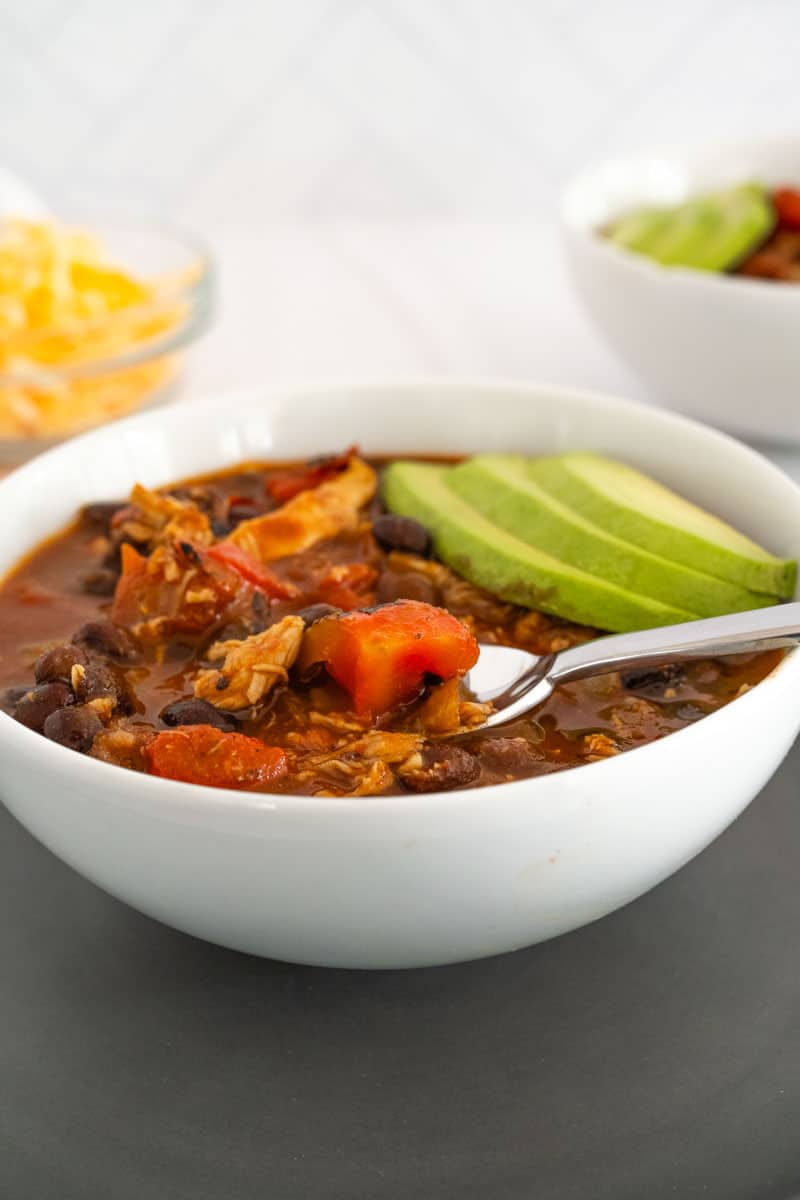 close up of a spoonful of chili in a white bowl with a chunk of tomato on spoon with sliced avocado on the side of the bowl. 
