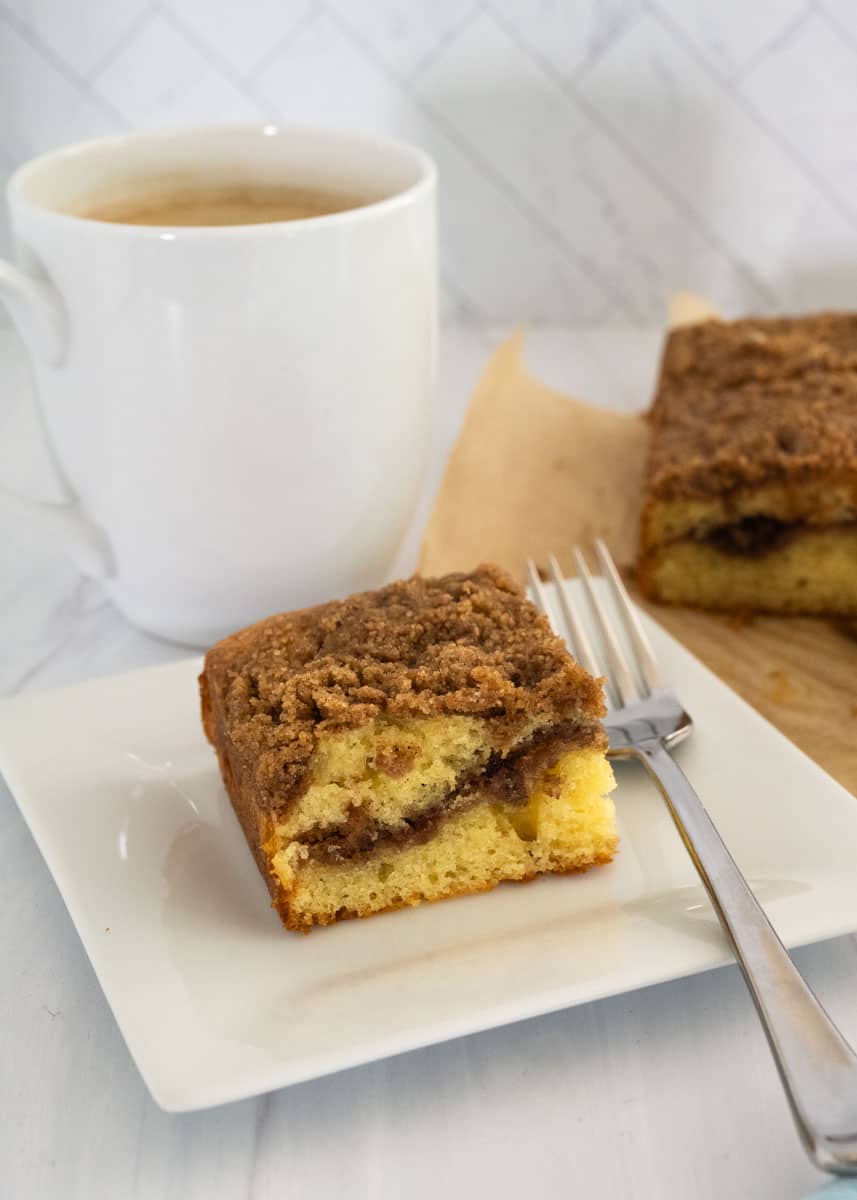 Close up picture of a slice of coffee cake next to a fork and white mug in the background. 