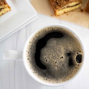 Re eye cup of coffee in white mug next to slices of coffee cake barely seen.