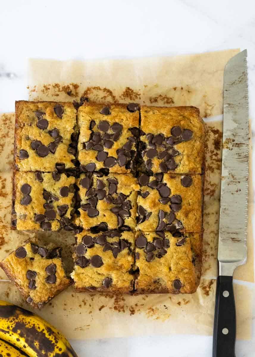 Aerial shot of anana snack cake cut into 9 pieces, with one piece bitten. A knife and ripe banana rest near the cake. 