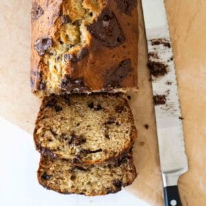 banana bread with chocolate chunks on a piece of parchment paper sliced. there is a knife next to the banana bread loaf.