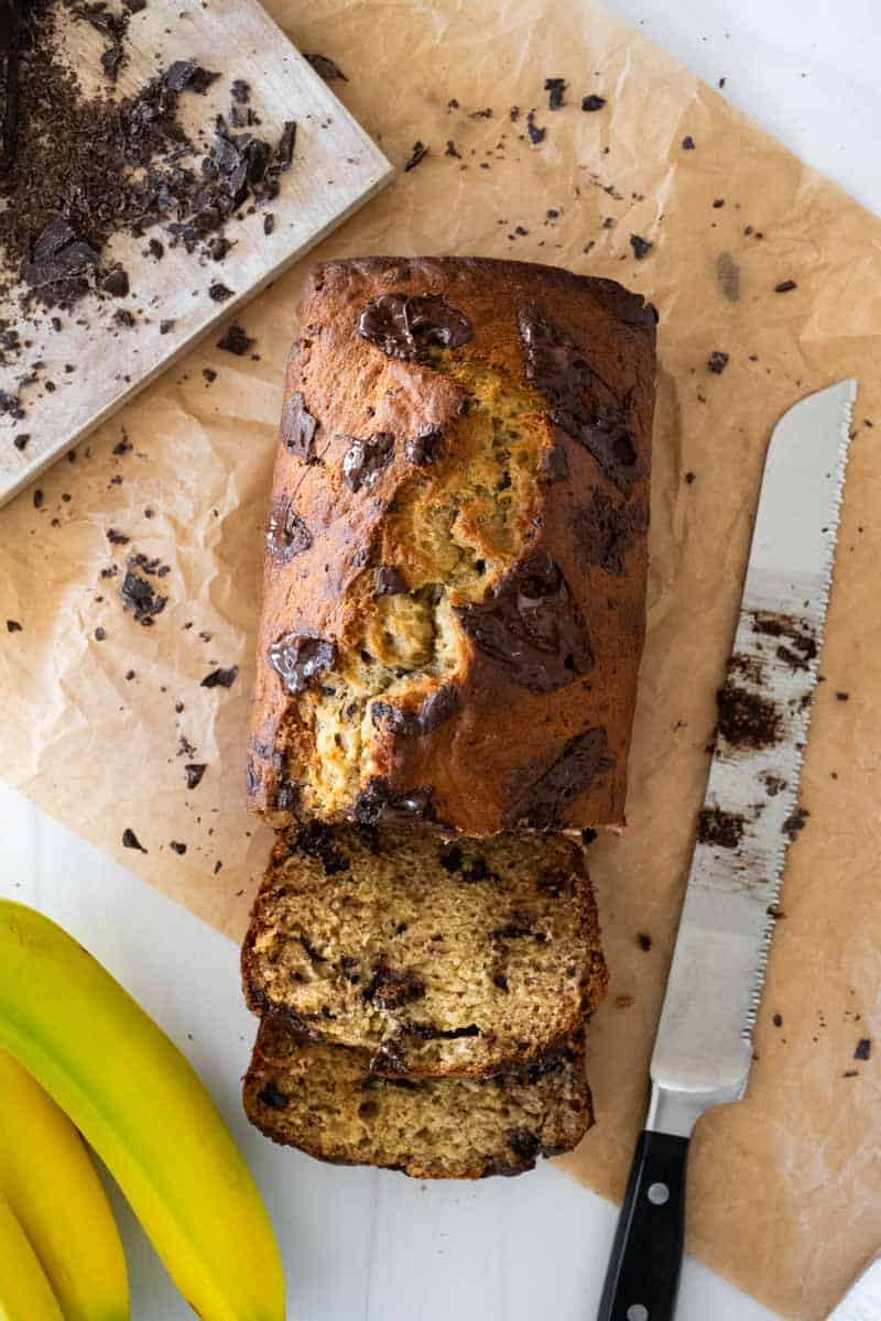 Flat lay picture of a sliced banana bread loaf with a chocolate-covered knife, bananas, and chopped chocolate around the loaf. 