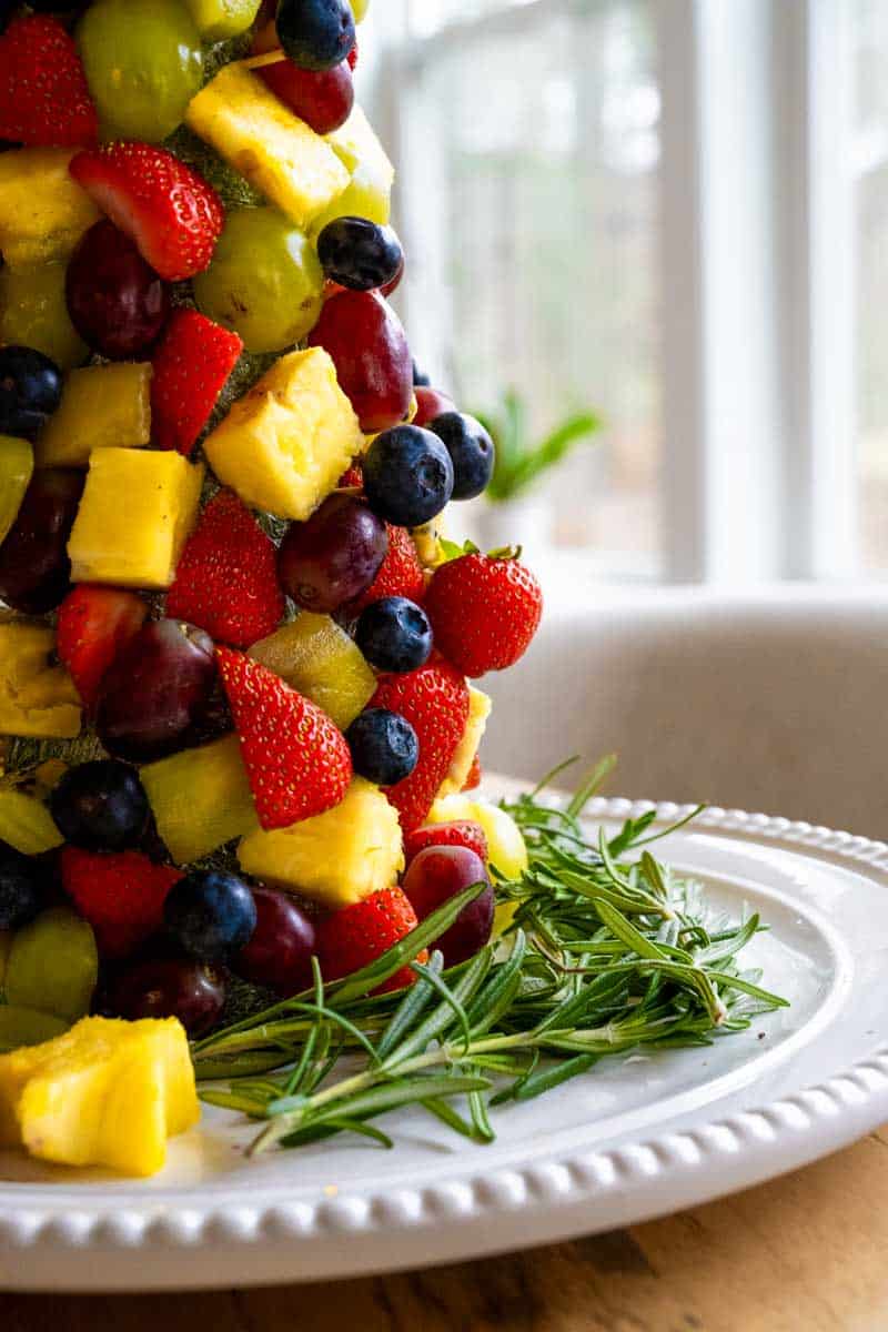 Up close picture of the side of the fruit Christmas tree with strawberries, kiwi, pineapple, and blueberries forming the tree garnished by fresh sprigs of rosemary. 