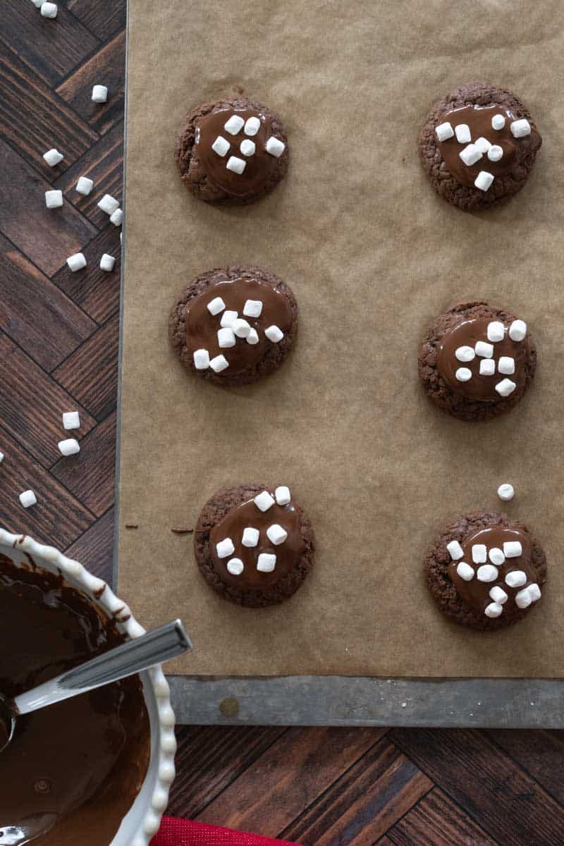 Six cookies on a parchment lined baking tray with mini marshmallows scattered around. 