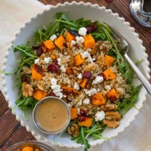 Pumpkin and quinoa salad in a ridged white bowl with arugula, pumpkin, quinoa, cranberries, goat cheese, and walnuts.