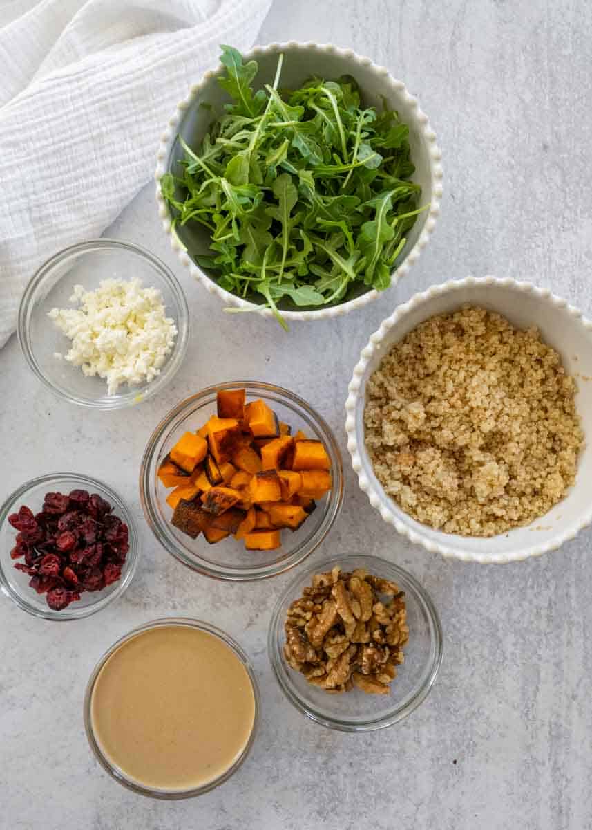 All of the ingredients needed to make the Pumpkin and Quinoa Salad in individual bowls, including arugula, quinoa, roasted pumpkin, goat cheese, walnuts, dried cranberries, and balsamic dressing. 