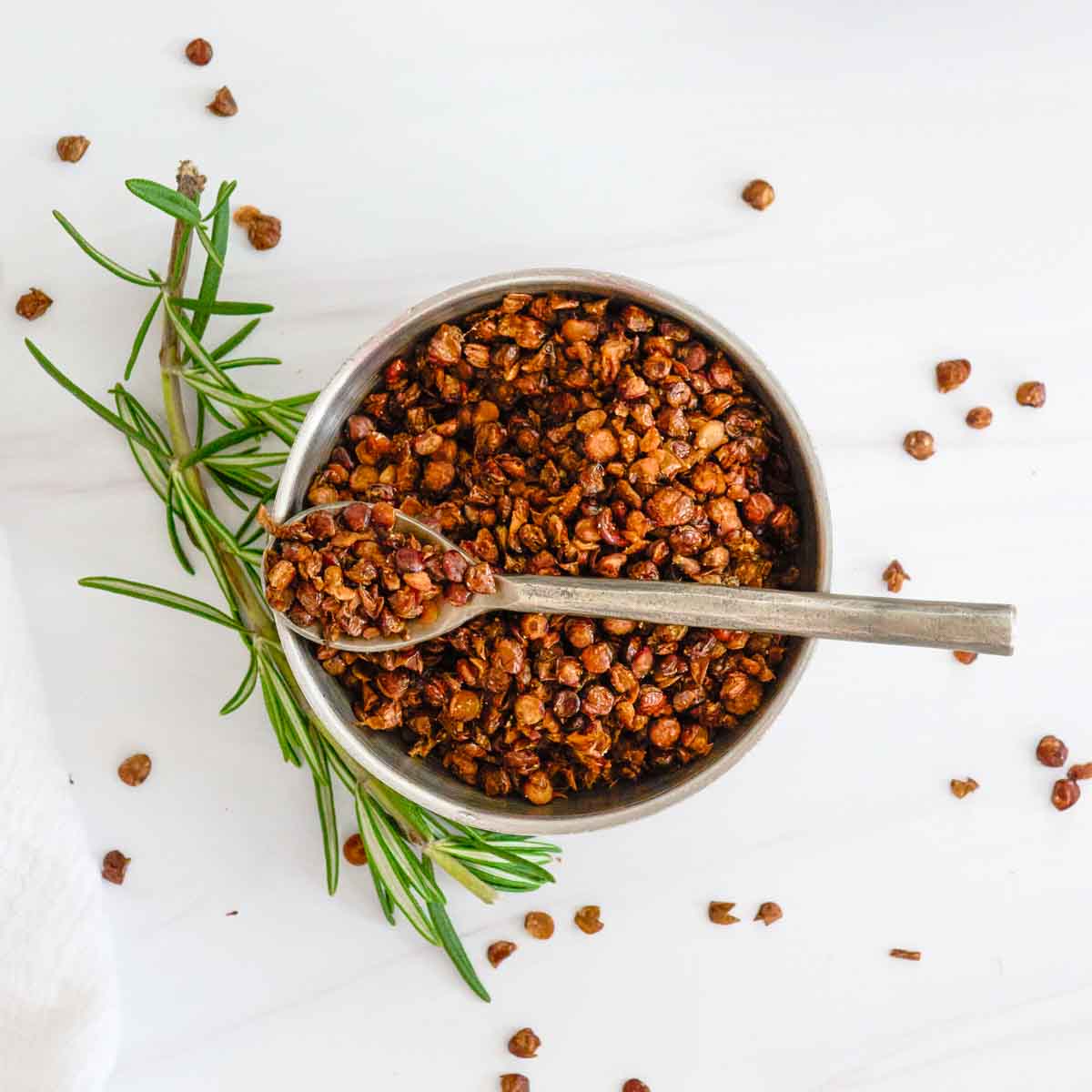 flat lay of white bowl of roasted lentils with a spoon full of lentils and a sprig of rosemary and loose lentils around the bowl.