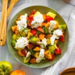 green plate with heirloom tomatoes, basil, and burrata surrounded by whole tomatoes and wooden salad spoons.