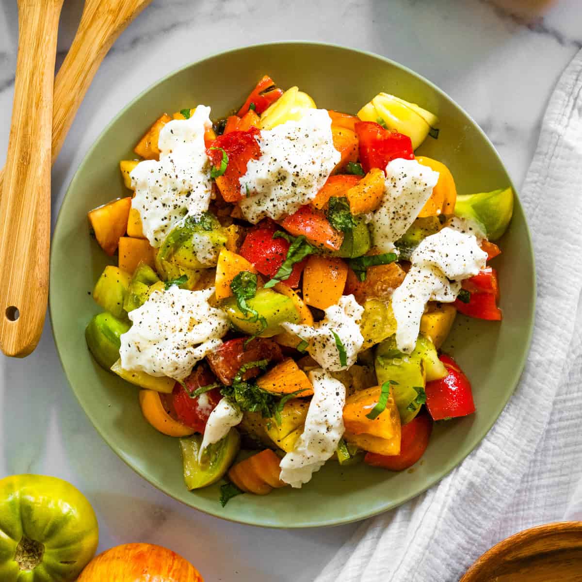 green plate with heirloom tomatoes, basil, and burrata surrounded by whole tomatoes and wooden salad spoons.