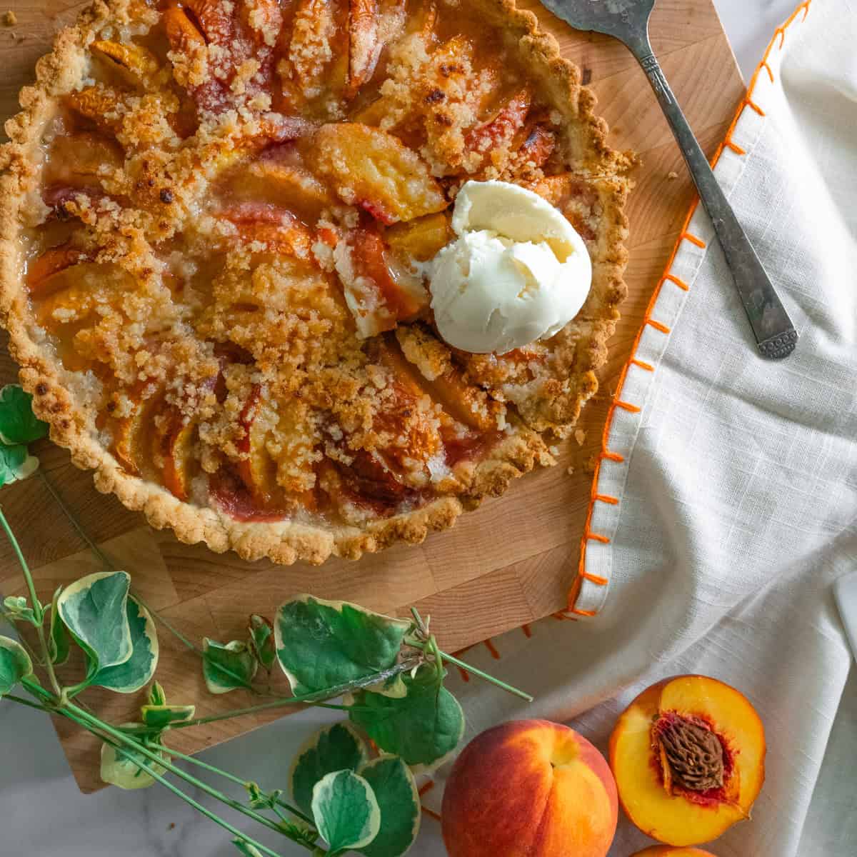 large peach tart on wooden cutting board with a scoop of icecream on one of the slices and peaches and green vinery around.