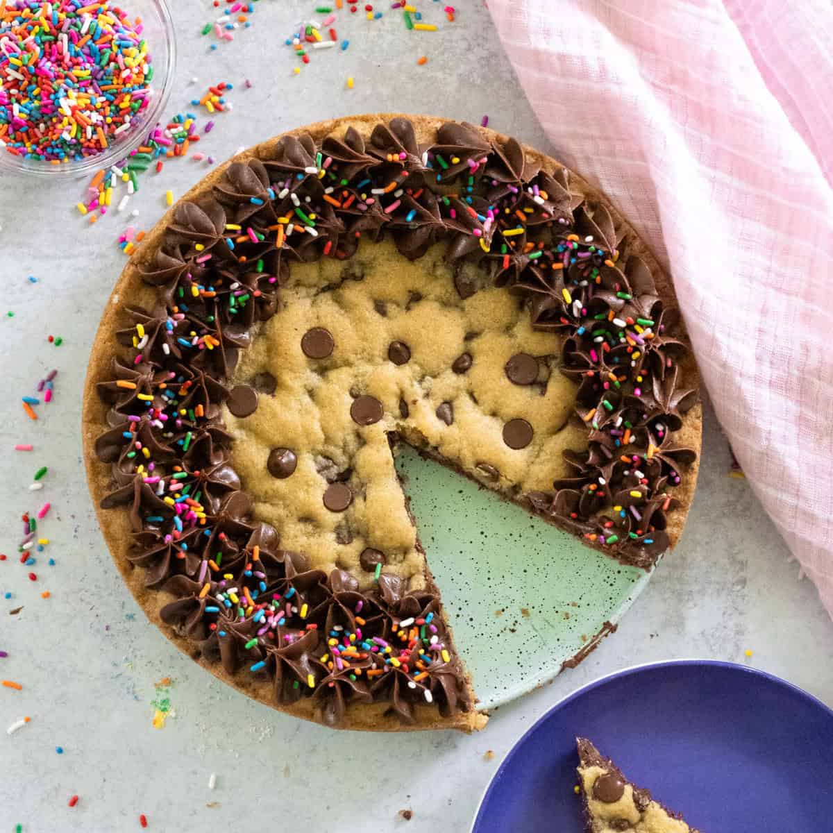 Aerial photo of chocolate chip cookie celebration cake with a slice cut out with sprinkles strewn about and a pink cloth on the counter.