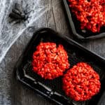 Picture of two red velvet rice krispie treats made to look like two raw meat burgers in a black tray.