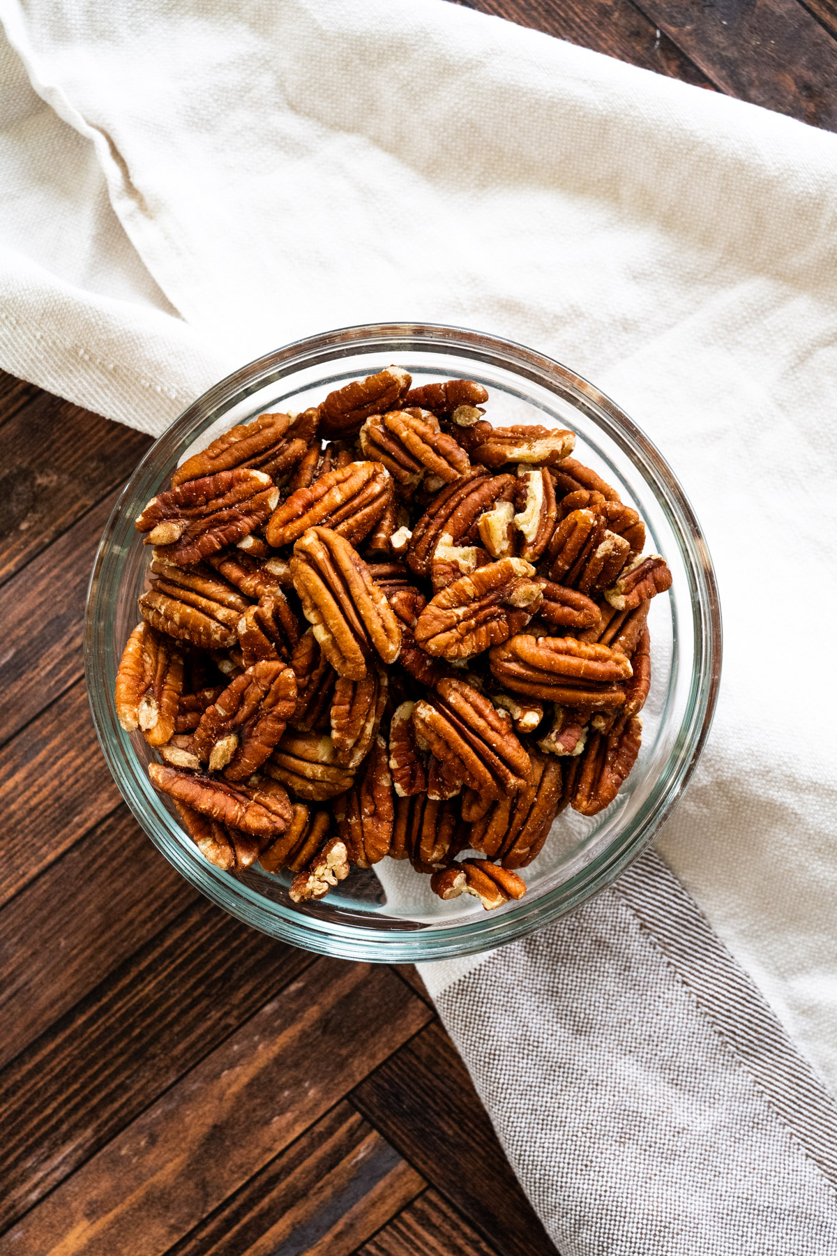Clear bowl full of raw pecan halves under a cream colored napkin against a wooden counter.
