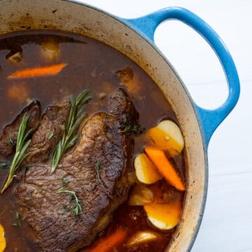 Blue Dutch oven with large chuck roast submerged in broth and carrots and potatoes.