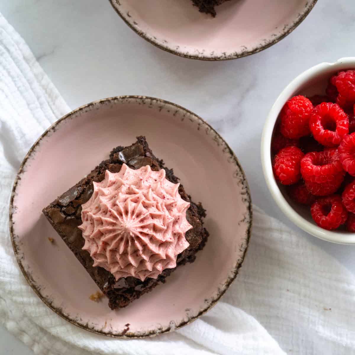 aerial of a pink plate with a square brownie on top and piped raspberry cream cheese frosting next to a bowl of raspberries.