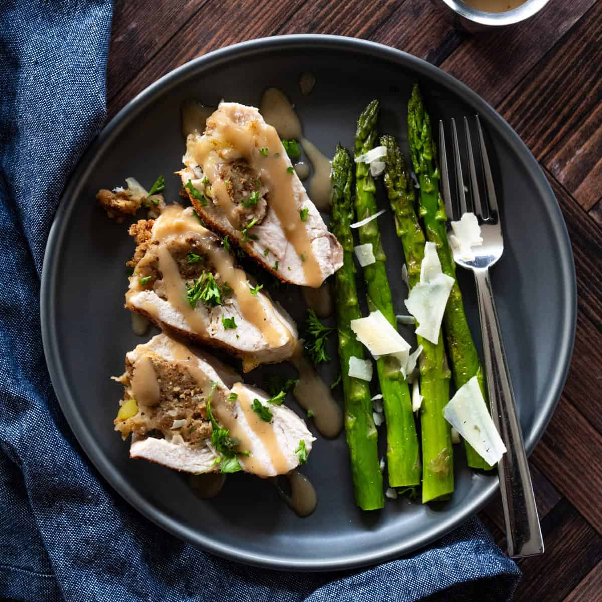 dark brown plate with three sliced pieces of chicken breast on their side to expose the stuffing that's inside the chicken breast. asparagus with parmesan cheese flakes is served on the side.