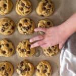 Small children's hand grabbing a chocolate chip cookie from a baking sheet with several other cookies.