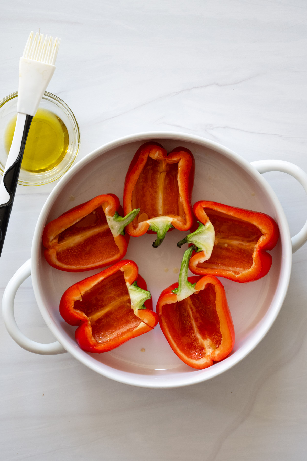 Step 1 of the directions showing red bell peppers sliced from top to bottom and placed in a round casserole dish. A bowl of olive oil and a pastry brush are next to dish. 