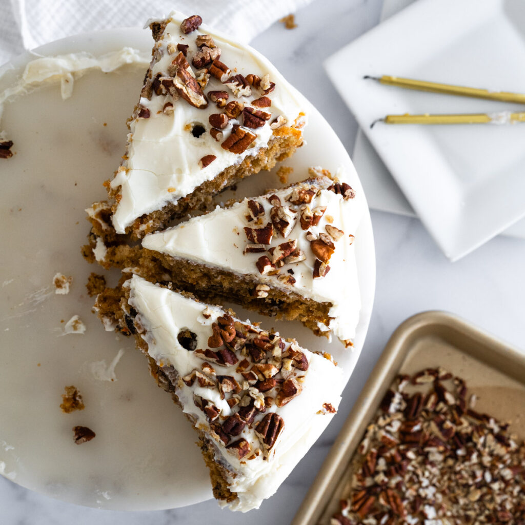 Aerial of three pieces of pecan carrot cake with a tray of toasted pecans and candles around the cake.