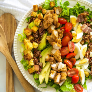 A large salad served on a white platter, including romaine lettuce, fried chicken tenders, hard boiled eggs, tomatoes, avocado, pecans, bacon, and cornbread croutons.
