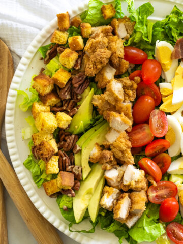 A large salad served on a white platter, including romaine lettuce, fried chicken tenders, hard boiled eggs, tomatoes, avocado, pecans, bacon, and cornbread croutons.