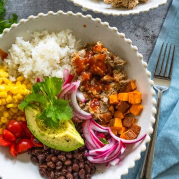 close up of a pulled pork bowl in a white scalloped bowl, including pulled pork with BBQ sauce, white rice, avocado, corn, tomatoes, black beans, pickled onions, and sweet potatoes.