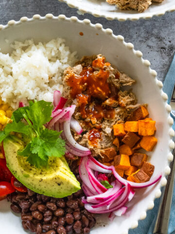 close up of a pulled pork bowl in a white scalloped bowl, including pulled pork with BBQ sauce, white rice, avocado, corn, tomatoes, black beans, pickled onions, and sweet potatoes.