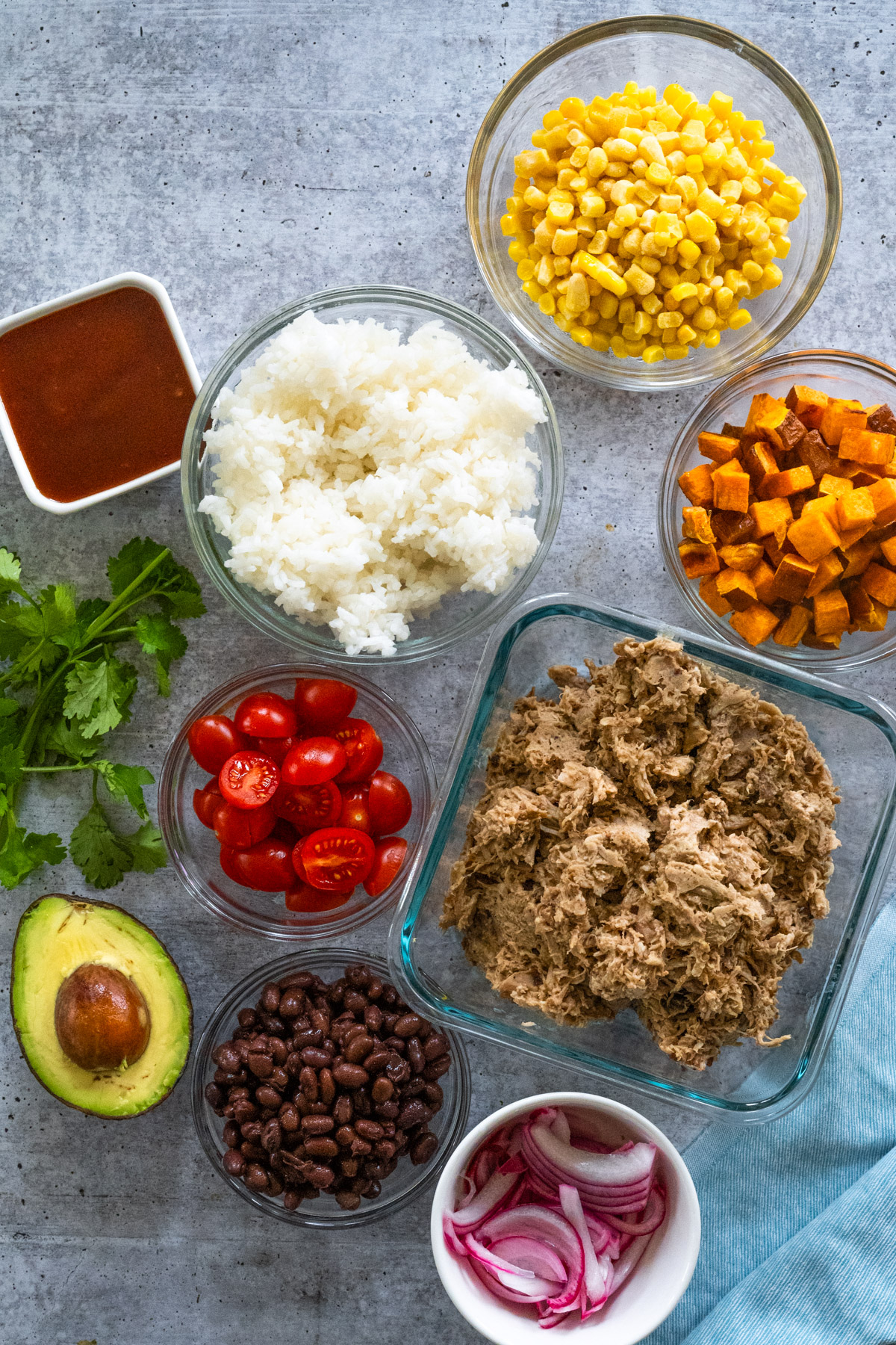 Ingredients needed to make a pulled pork bowl, including pulled pork, sweet potatoes, rice, BBQ sauce, tomatoes, avocado, cilantro, black beans, and pickled onions. 