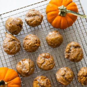 glazed pumpkin donut holes cooling on a wire rack against a white counter with two miniature pumpkins on the rack as well.