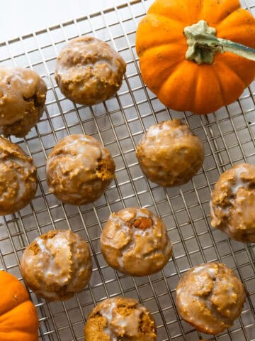 glazed pumpkin donut holes cooling on a wire rack against a white counter with two miniature pumpkins on the rack as well.