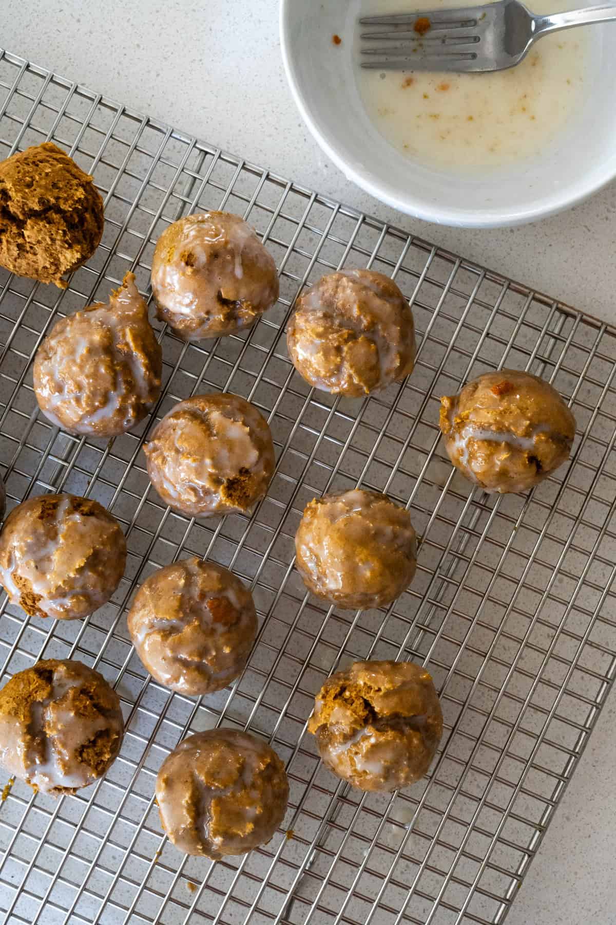 Glazed pumpkin donut holes cooling on a wire rack with a bowl of glaze in the upper-right corner of the frame.